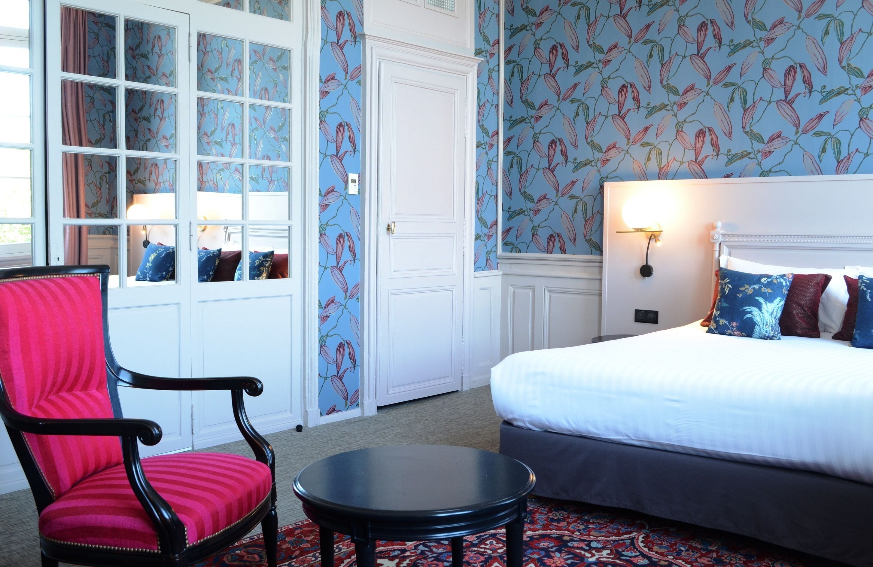 A hotel room with a bed, a red upholstered chair, a small table, and a window with curtains.