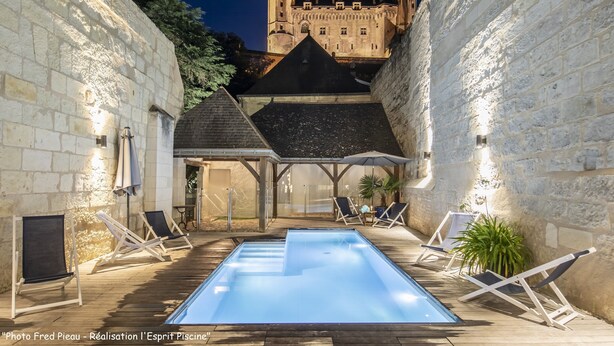 Una piscina con sillones de descanso y un área de estar cubierta, rodeada de muros de piedra y un castillo al fondo.