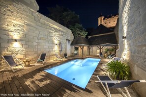 Una piscina con sillones de descanso y un muro de piedra, rodeada por un edificio de estilo castillo.
