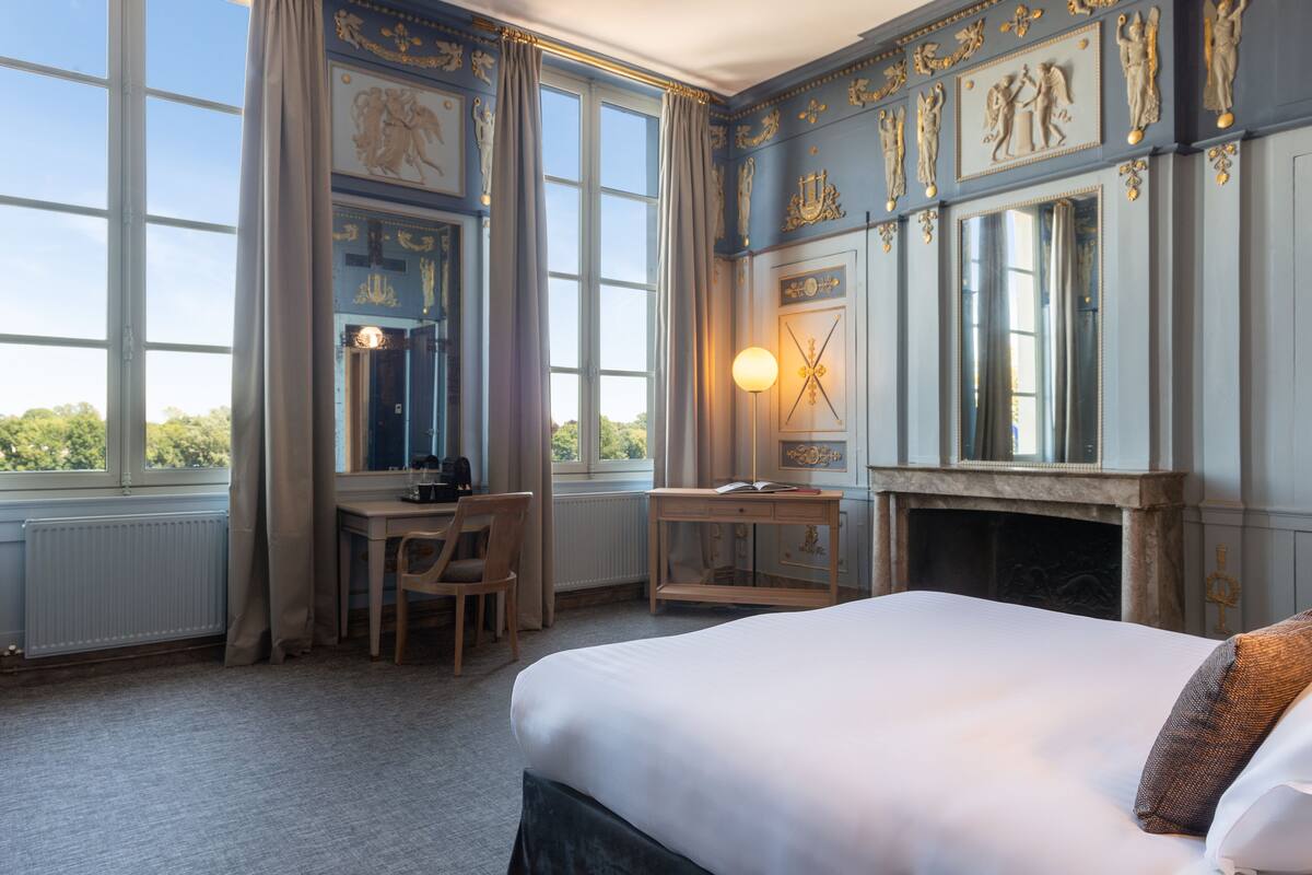 A hotel room with a large bed, a desk, a chair, a fireplace, and large windows.