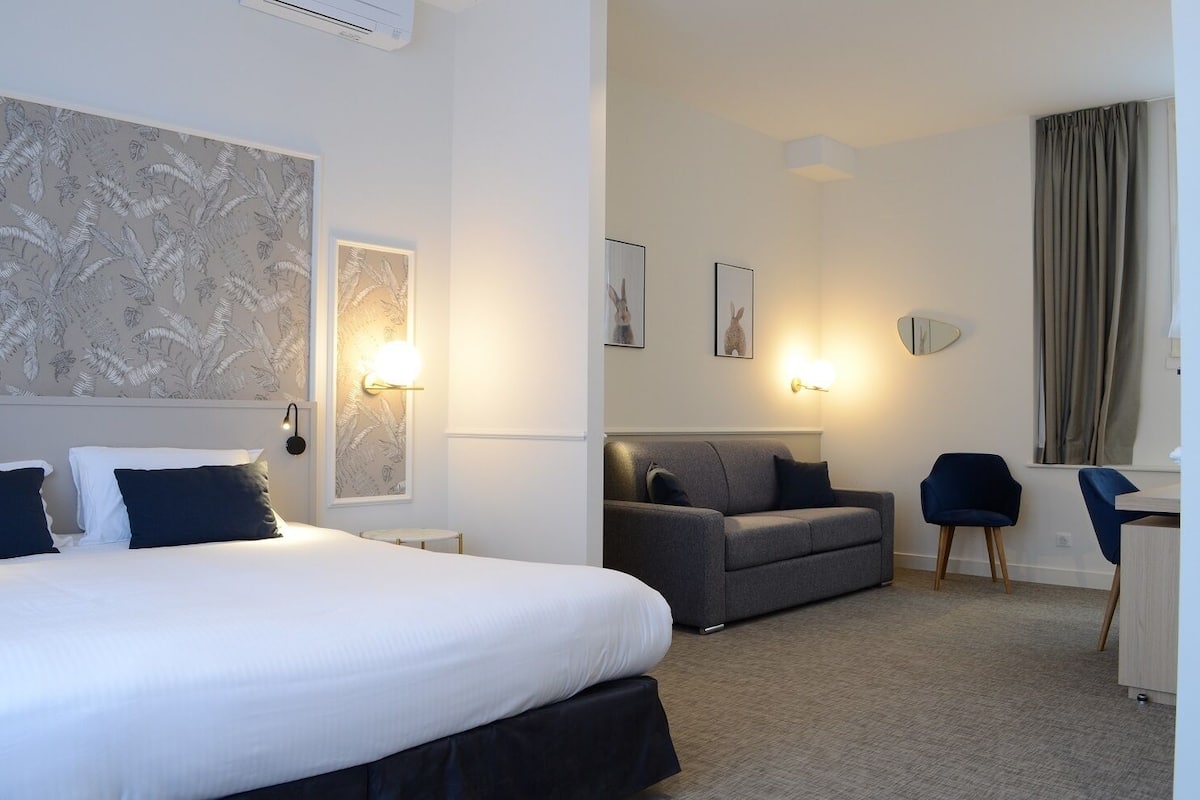 A hotel room with a large bed, a sofa, a desk, and a chair. There are wall-mounted lights and framed pictures on the walls.