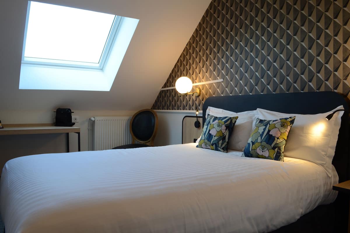 A bedroom with a skylight, a bed with white bedding, a round mirror, and a wall with a geometric pattern.
