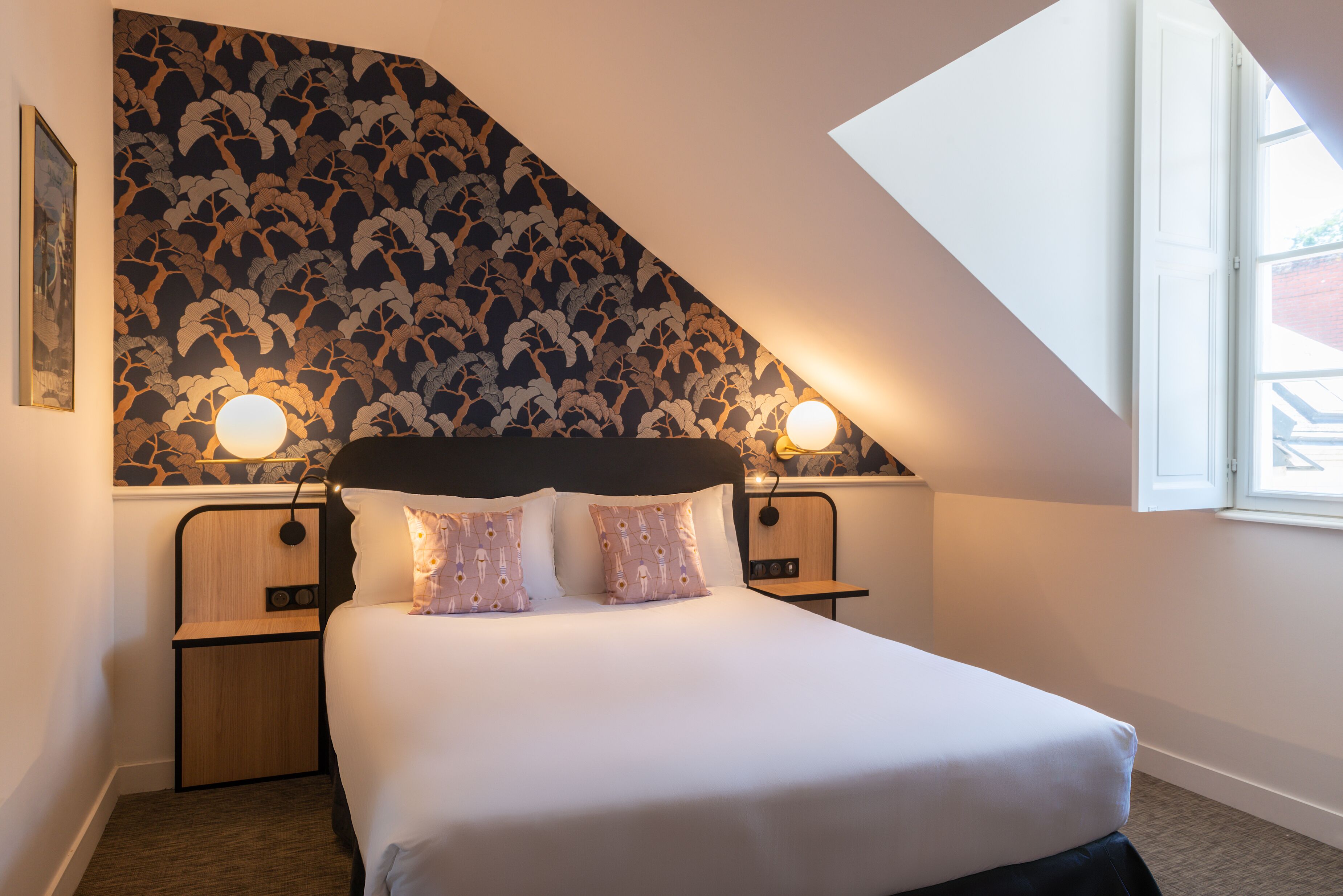 A bedroom with a sloped ceiling, a bed with white linens, two bedside tables with lamps, and a patterned wallpaper.