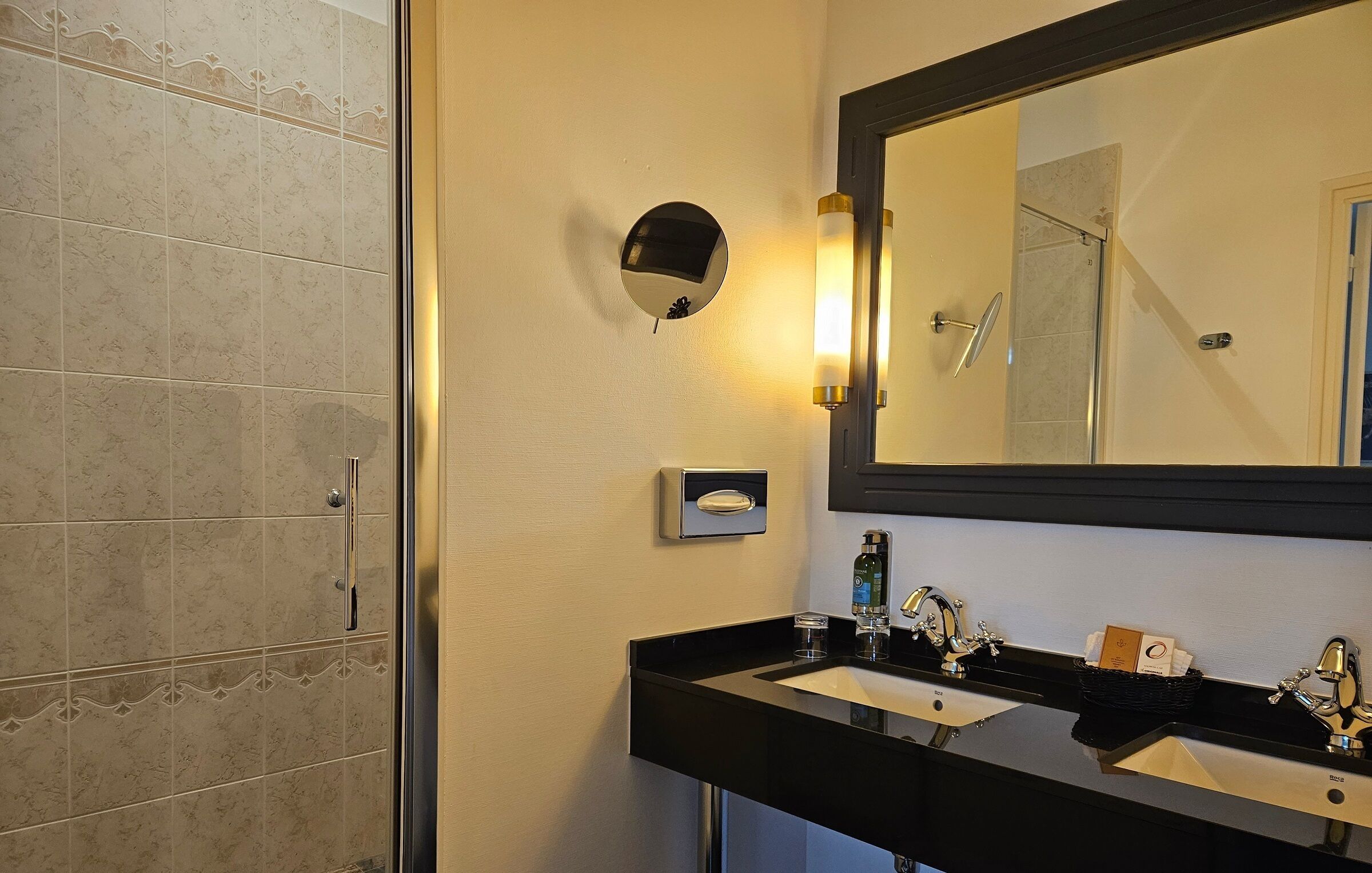Un baño con ducha, un lavabo doble y un espejo, además de una lámpara de pared.