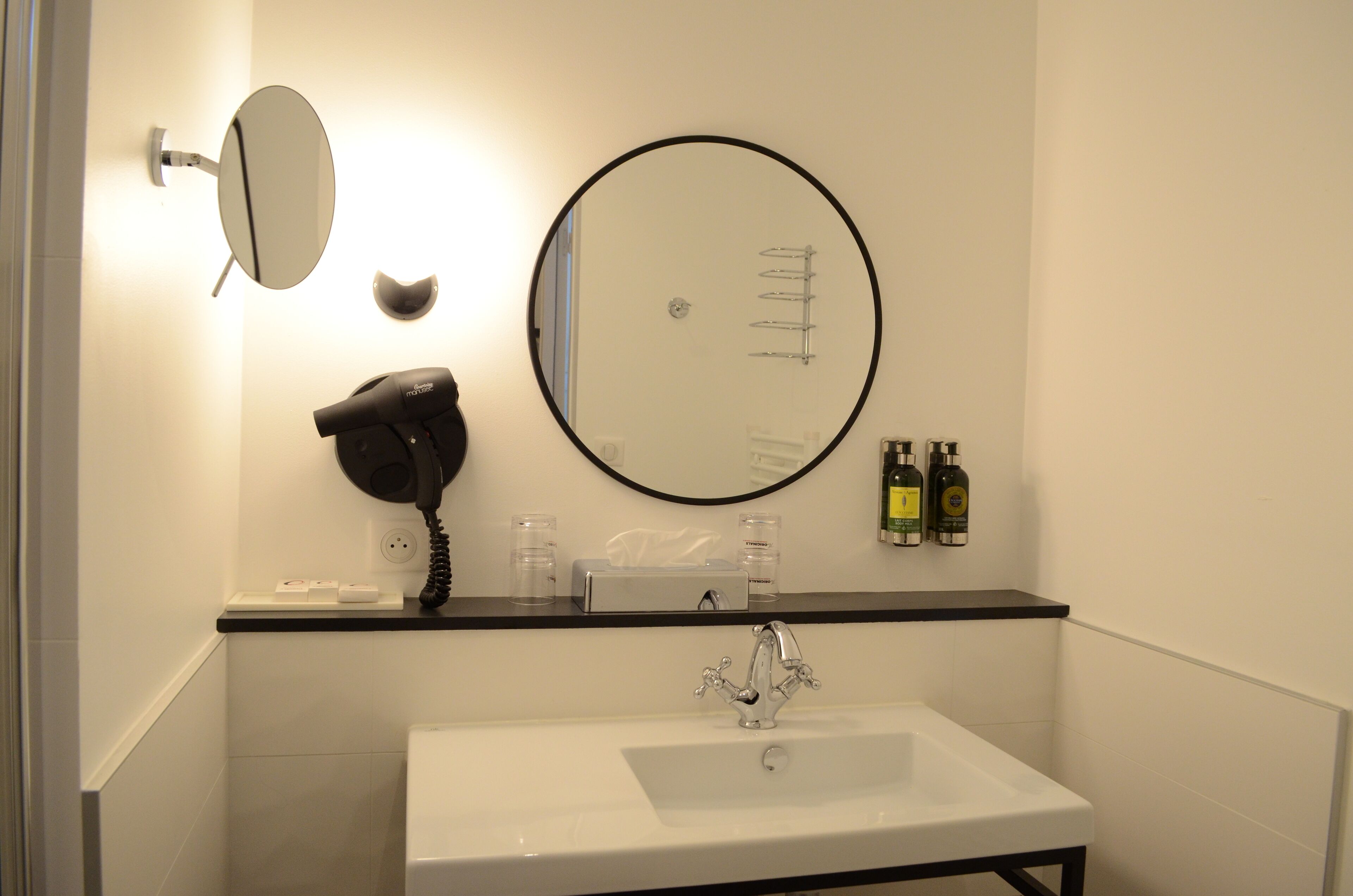 A bathroom with a white sink, a round mirror, a hairdryer, and toiletries on the countertop.