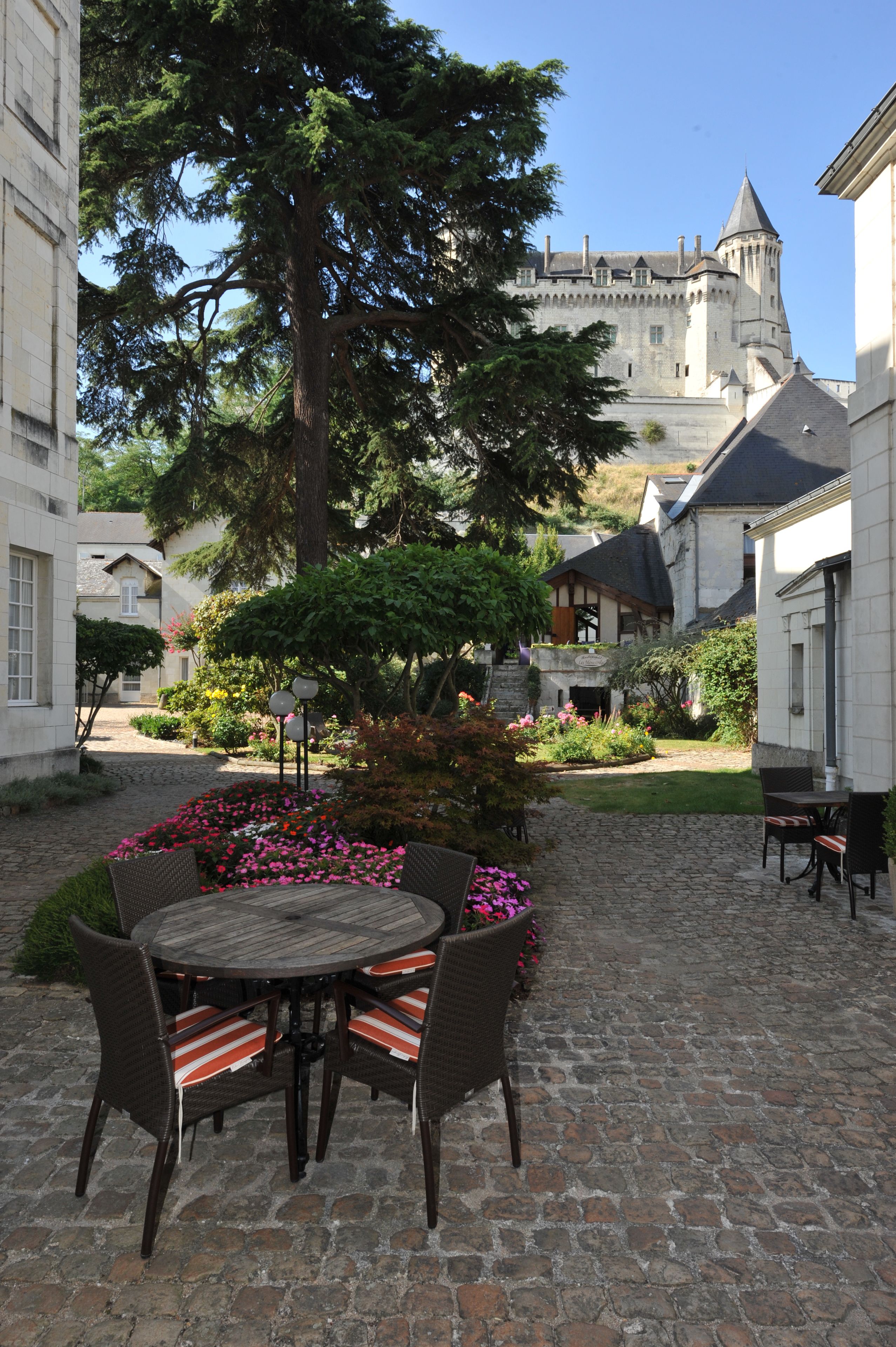 Un patio con una mesa y sillas de piedra, rodeado de flores y un árbol grande.