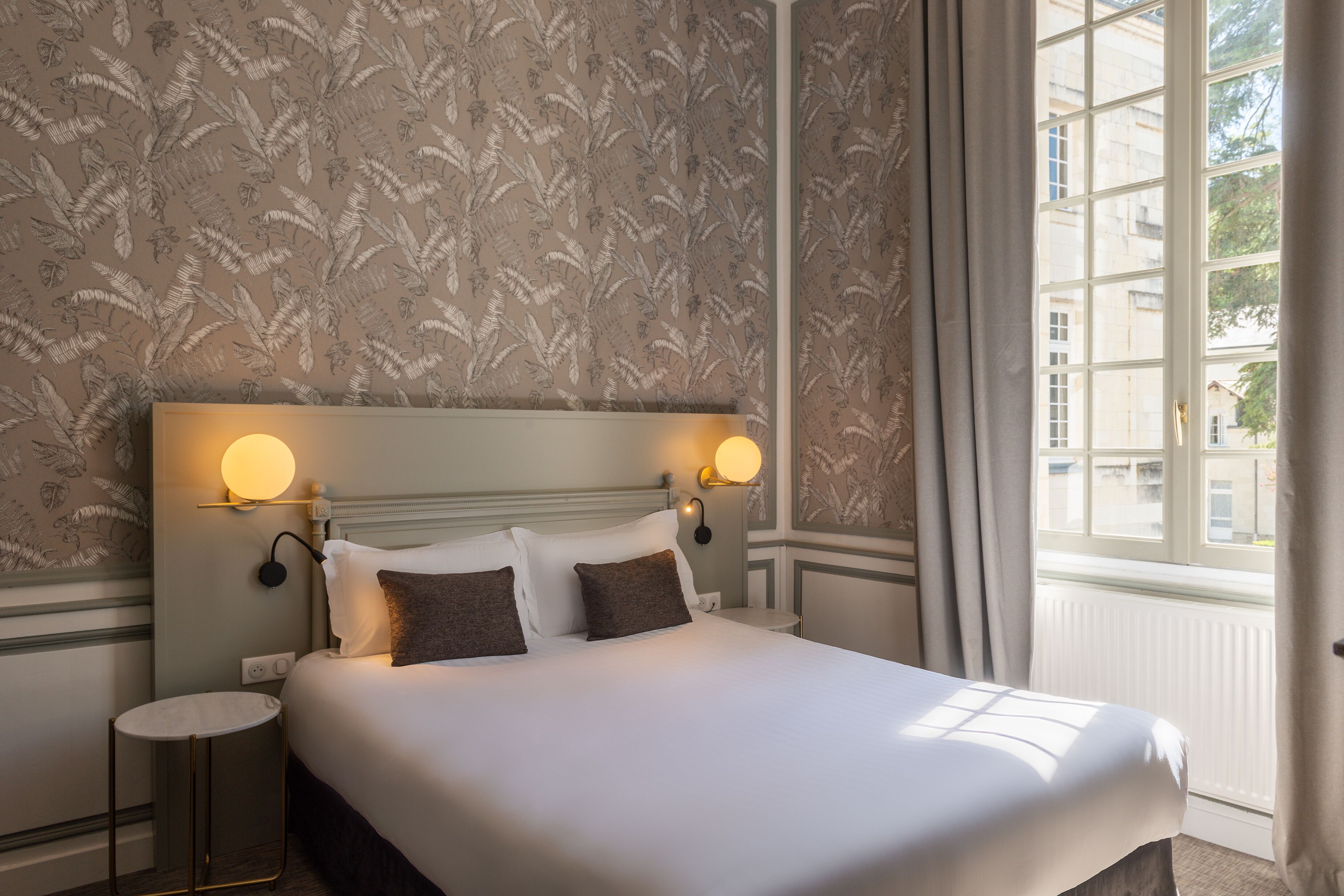 A neatly made bed with white and brown bedding, a round bedside table, and wall-mounted reading lamps.