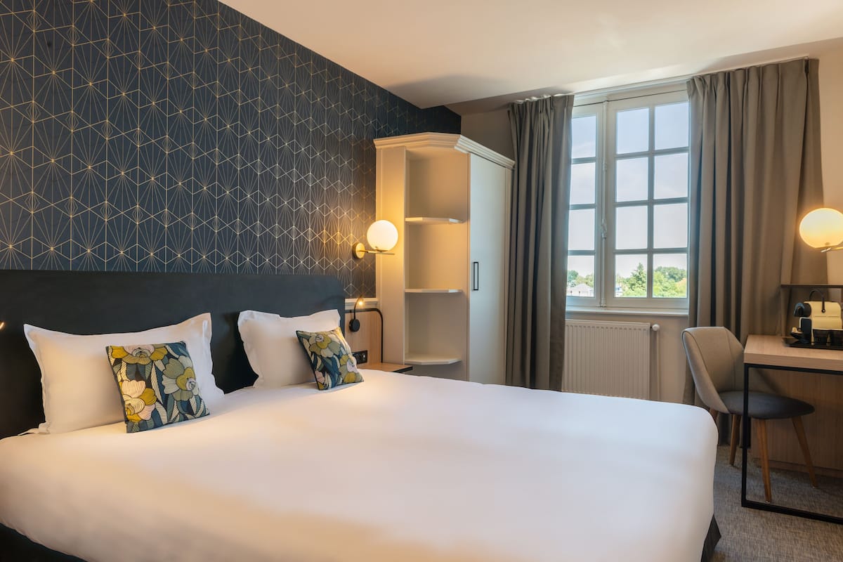 A hotel room with a large bed, a desk with a chair, a wardrobe, and a window with curtains.