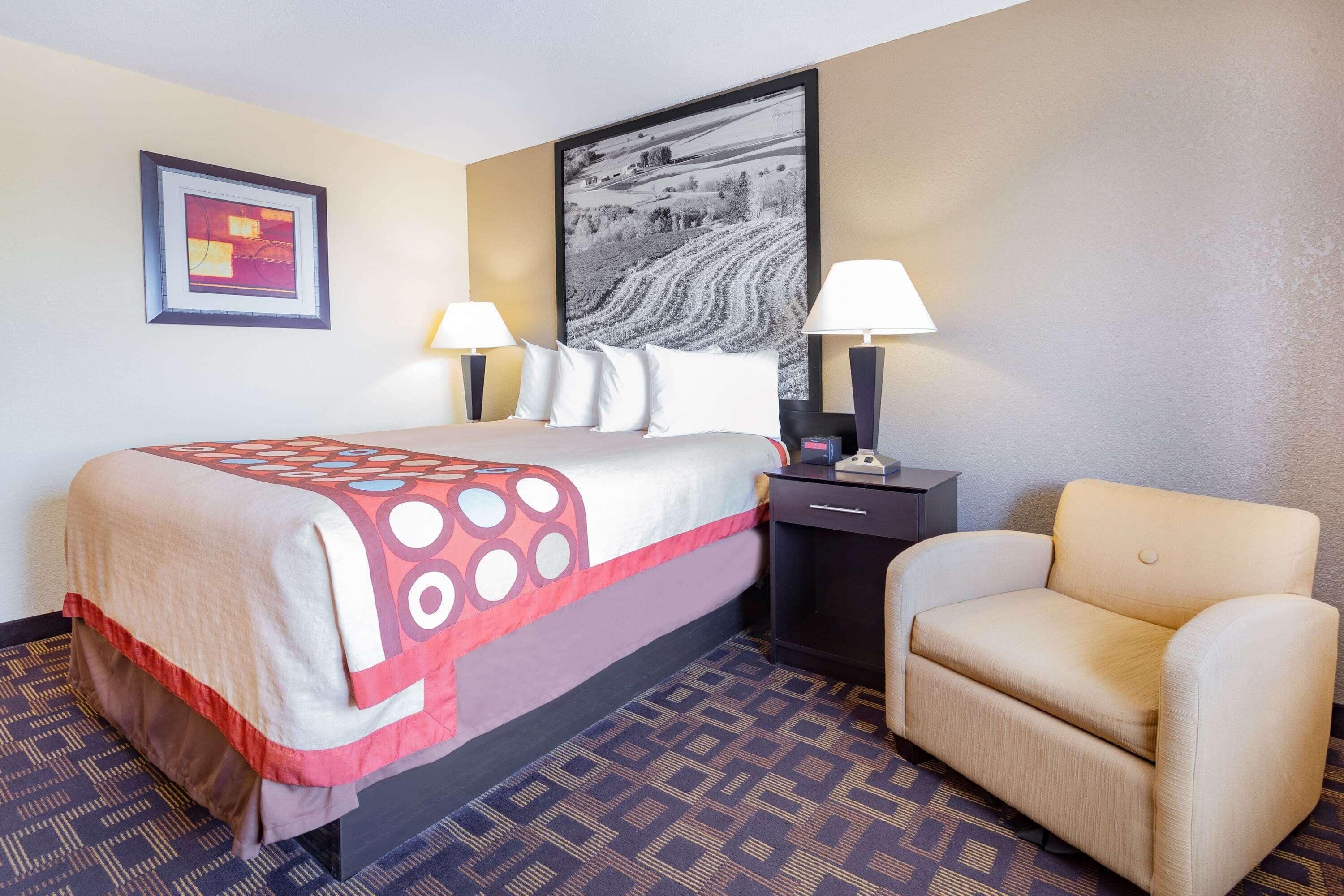 A hotel room with a bed, two bedside lamps, a chair, a nightstand, a framed picture, and a patterned carpet.