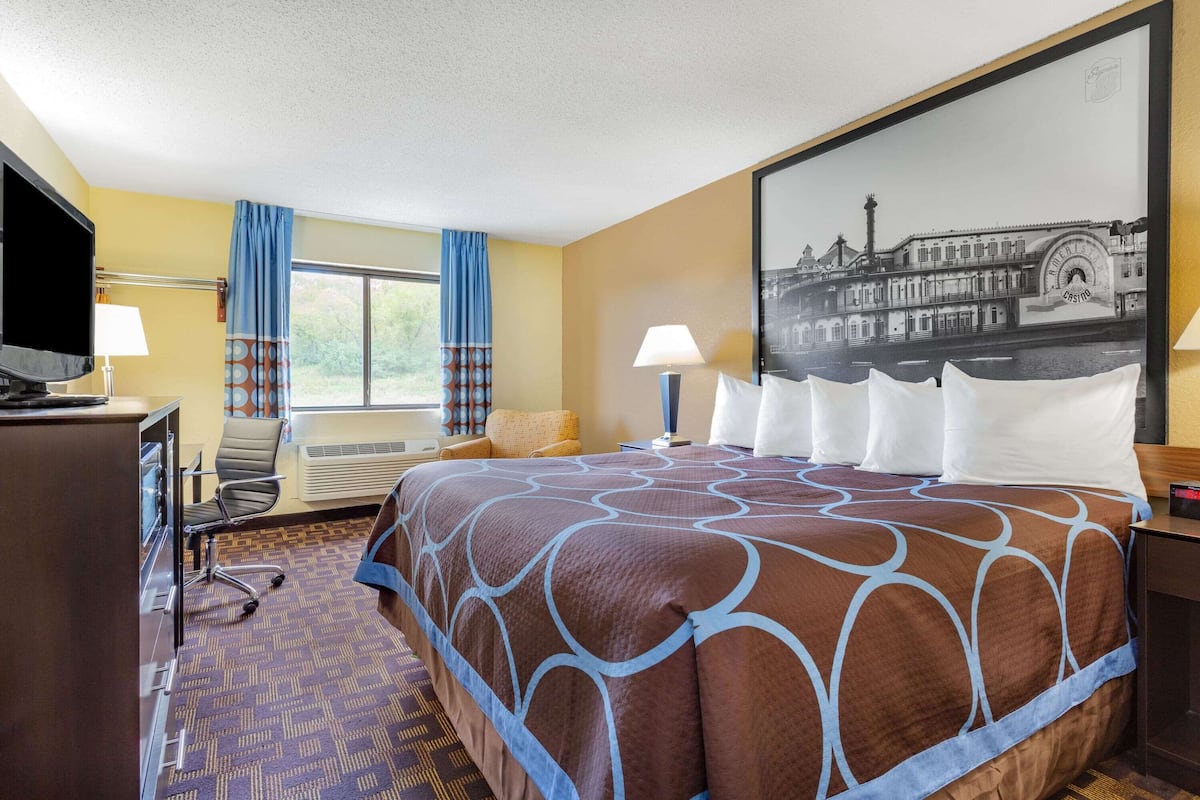 A hotel room with a large bed, a desk with a computer, a chair, a window with curtains, and a wall-mounted TV.