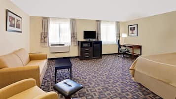 A hotel room with a sofa, a bed, a desk with a computer, a television, and an air conditioning unit.