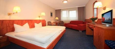Quarto de hotel com uma cama grande, uma escrivaninha com televisão, duas cadeiras e um sofá vermelho.