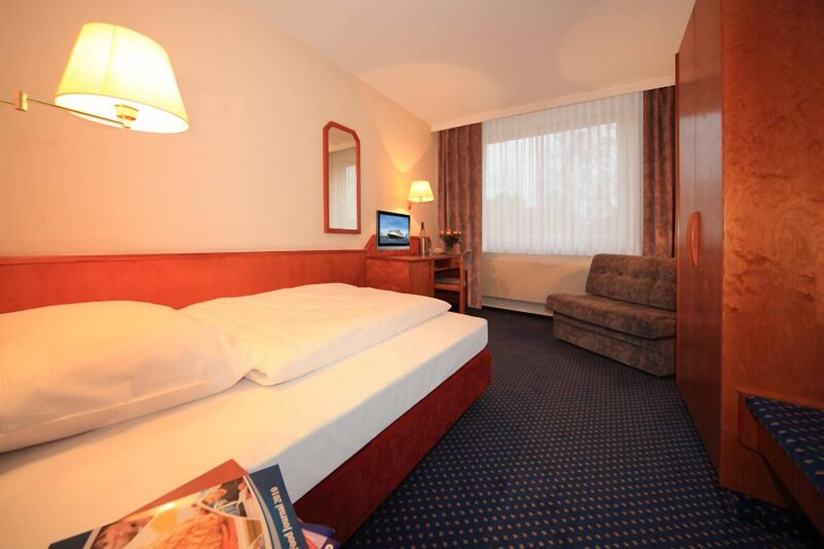 A hotel room with a large bed, a desk with a computer, a chair, a window with curtains, and a wooden wardrobe.