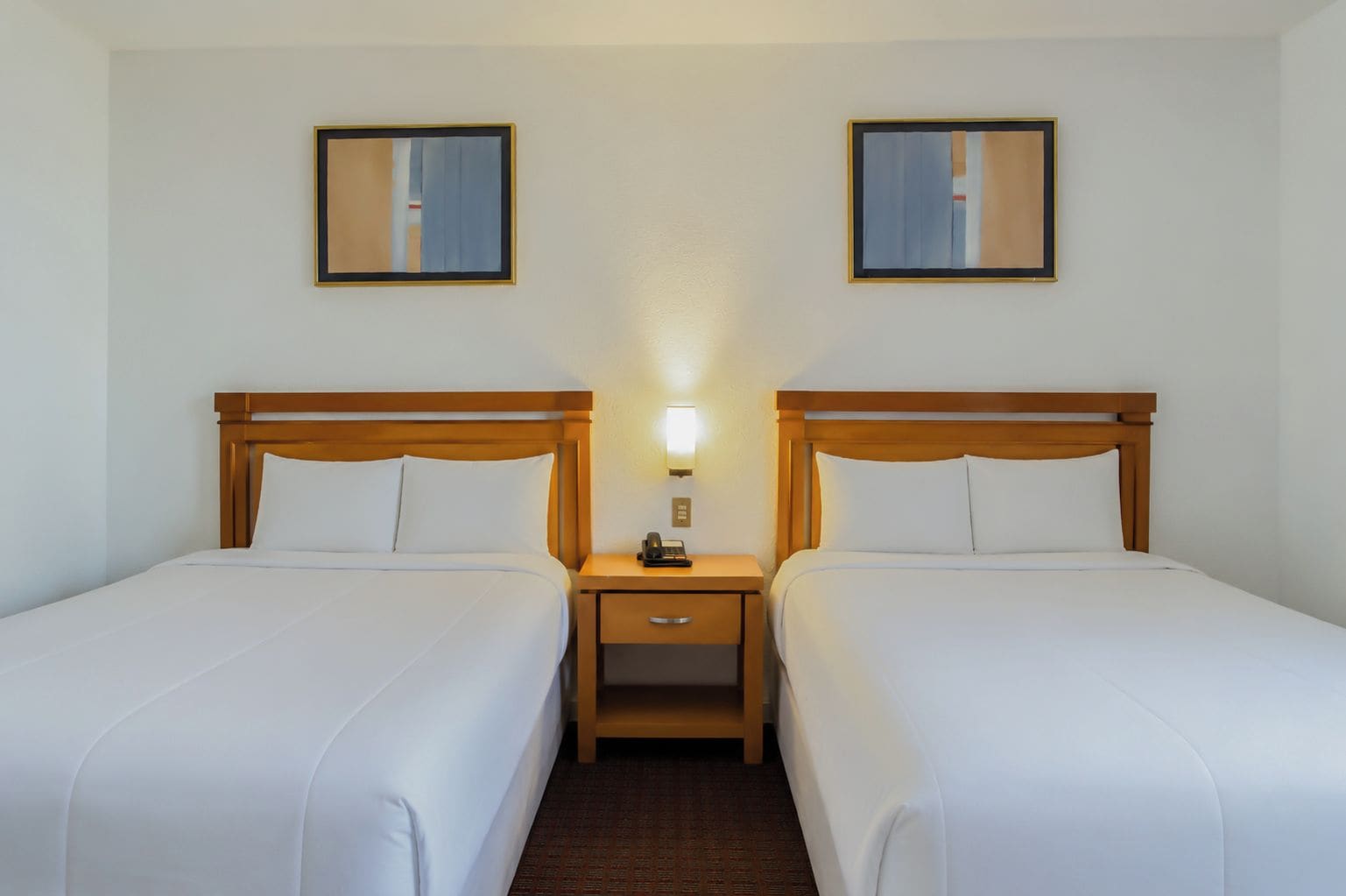 A hotel room with two single beds, a nightstand with a lamp, and two framed pictures on the wall.