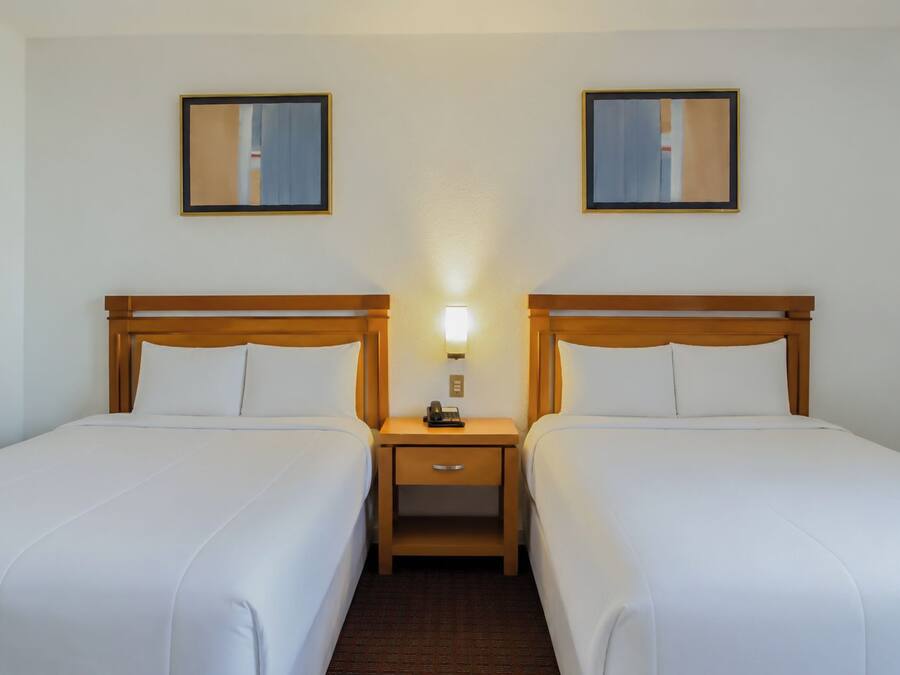 A hotel room with two single beds, a nightstand with a lamp, and two framed pictures on the wall.