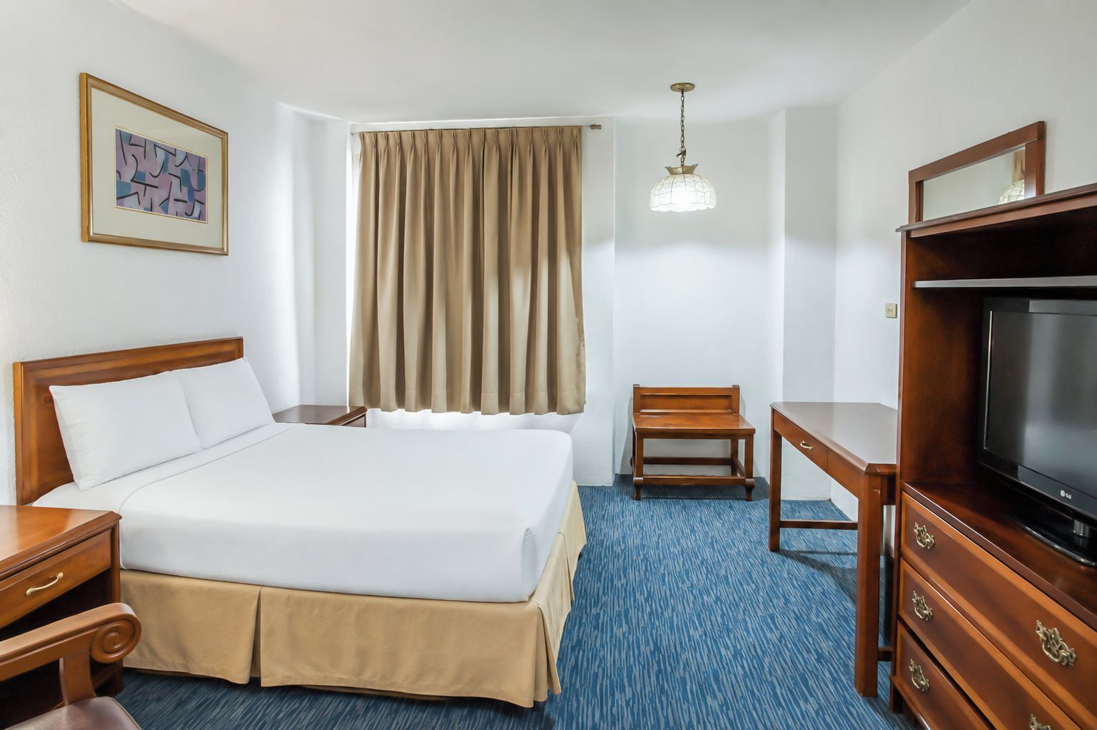 A hotel room with a bed, a desk, a television, a chair, a curtain, and a hanging light fixture.