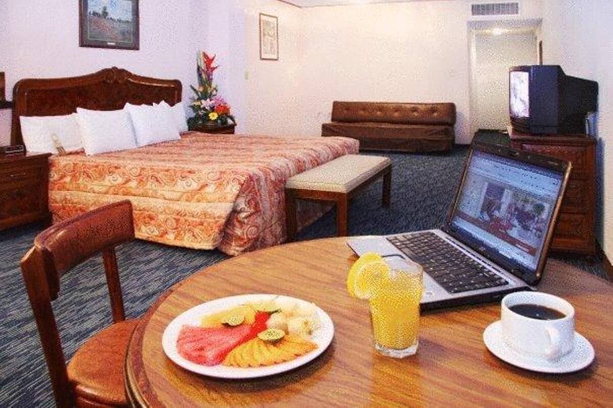A hotel room with a bed, a laptop, a plate of fruit, a glass of juice, and a cup of coffee on a wooden table.
