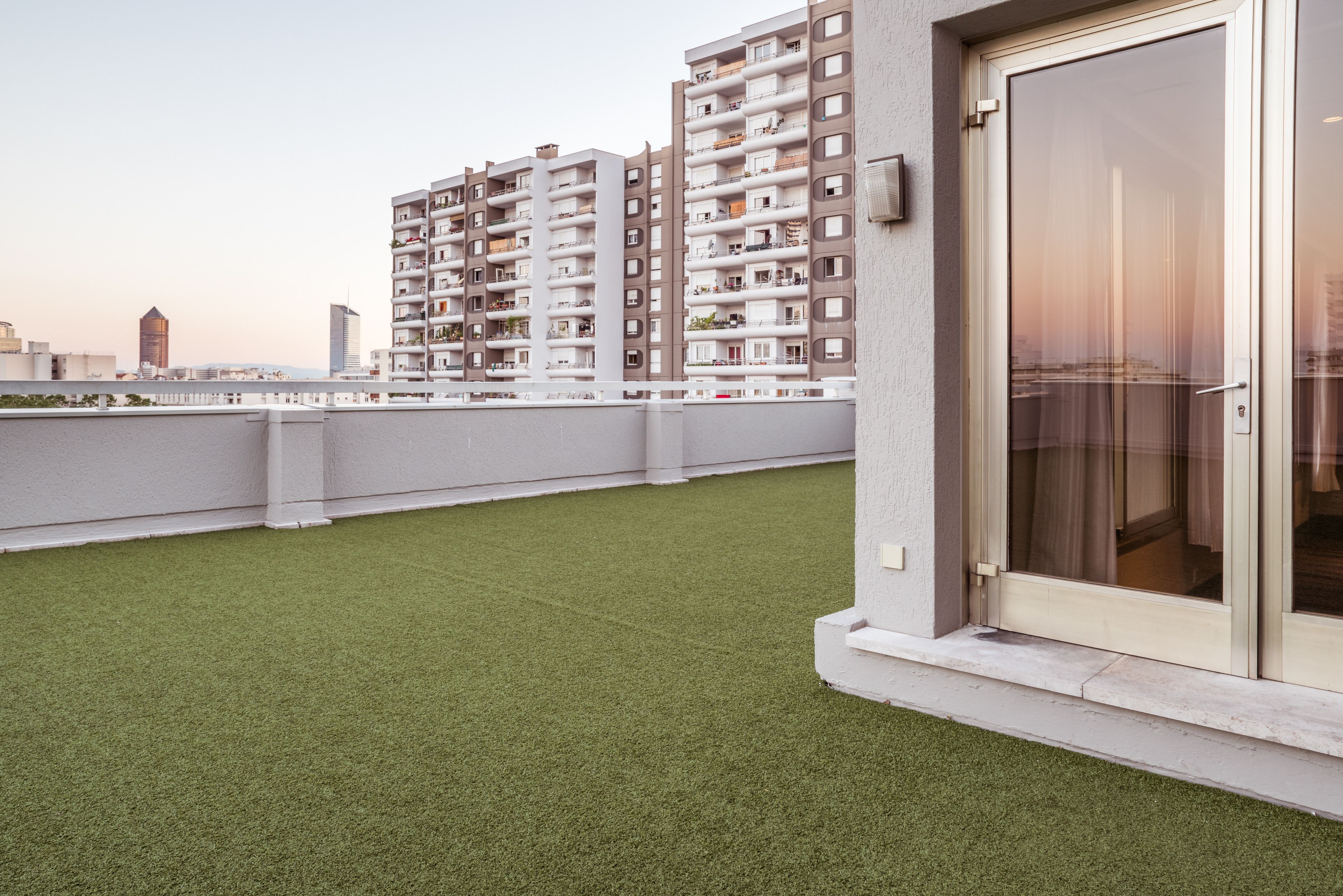 A rooftop terrace with artificial grass, a sliding glass door, and a view of high-rise residential buildings.