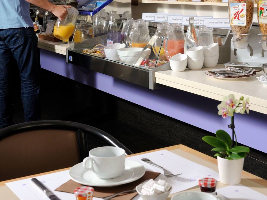 Um buffet de café da manhã com diversos alimentos e bebidas, um homem servindo suco de laranja e uma mesa posta com croissants e xícaras de café.