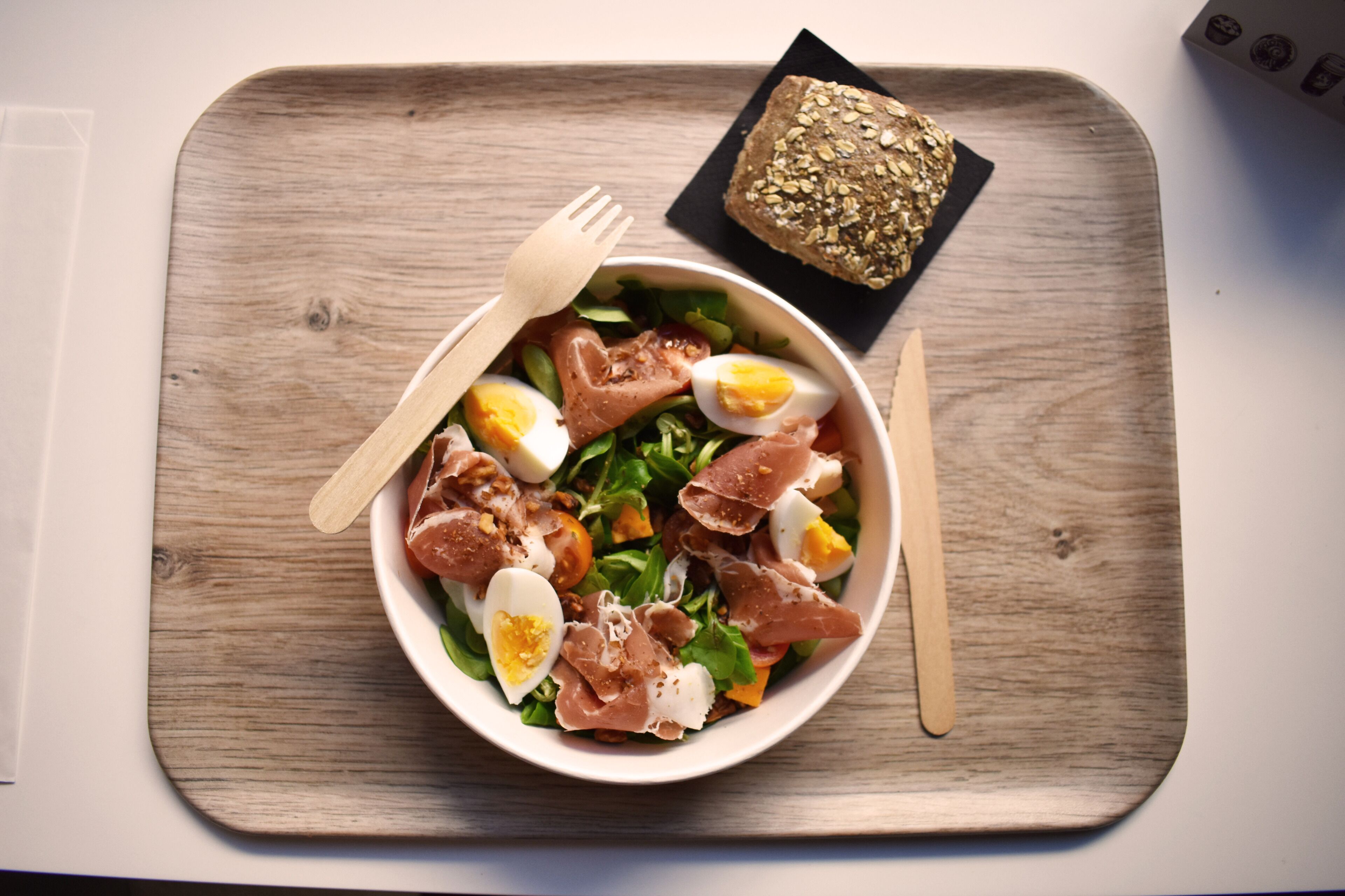 Een houten dienblad met een saladeschaal gevuld met sla, eieren en vlees, samen met een sneetje brood en een vork.