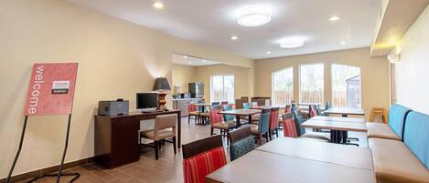 A hotel dining area with multiple tables and chairs, a reception desk, and a 'Welcome' sign.