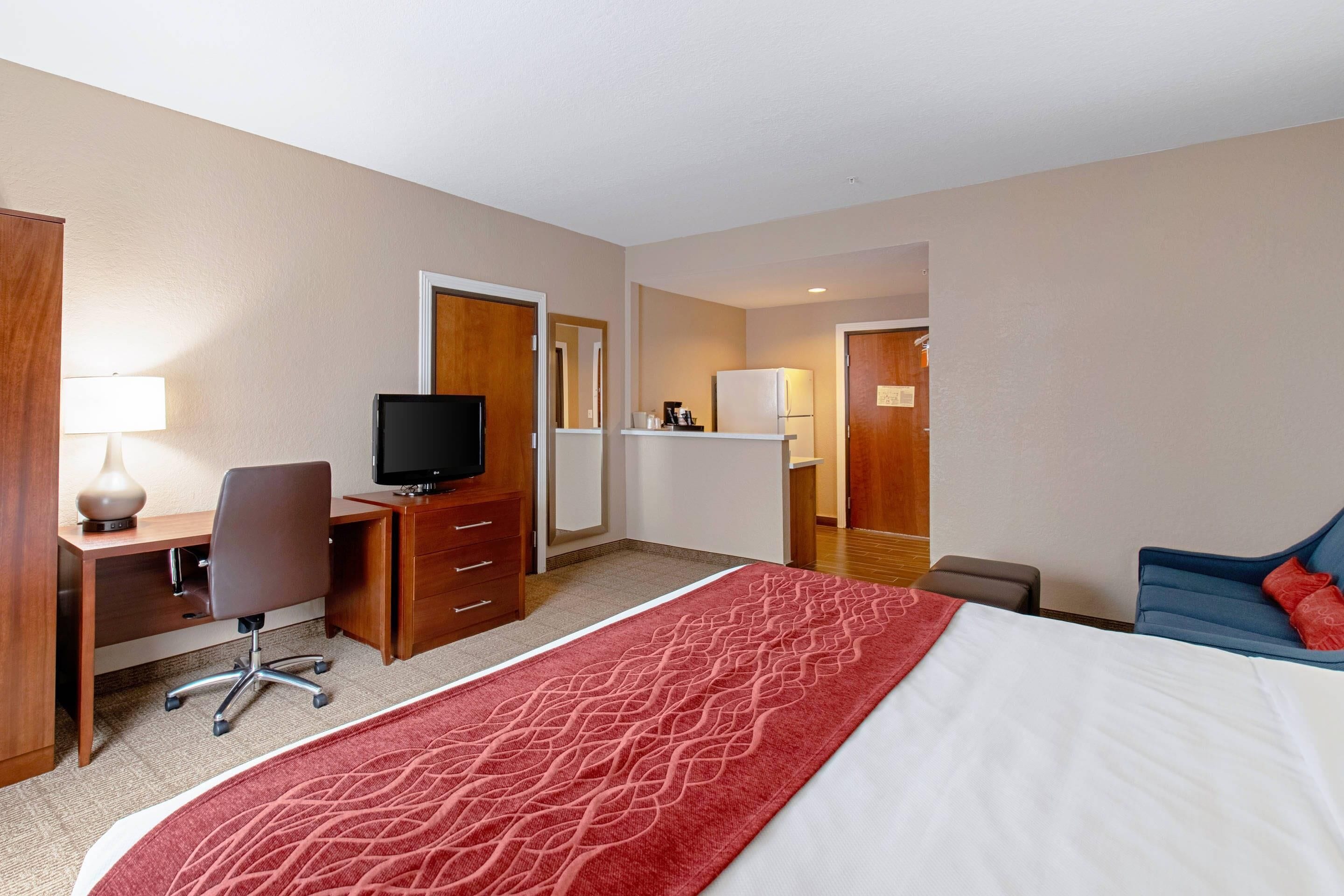 A hotel room with a bed, a desk with a computer, a television, a small kitchenette, and a seating area.