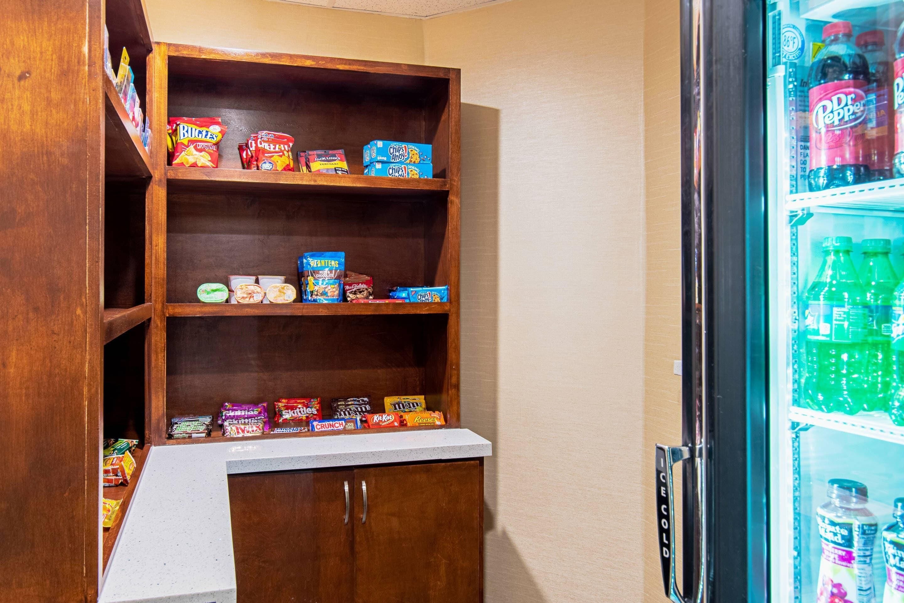 A kitchenette with a wooden shelf unit stocked with snacks and a refrigerator filled with beverages.