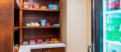 A kitchenette with a wooden shelf unit stocked with snacks and a refrigerator filled with beverages.