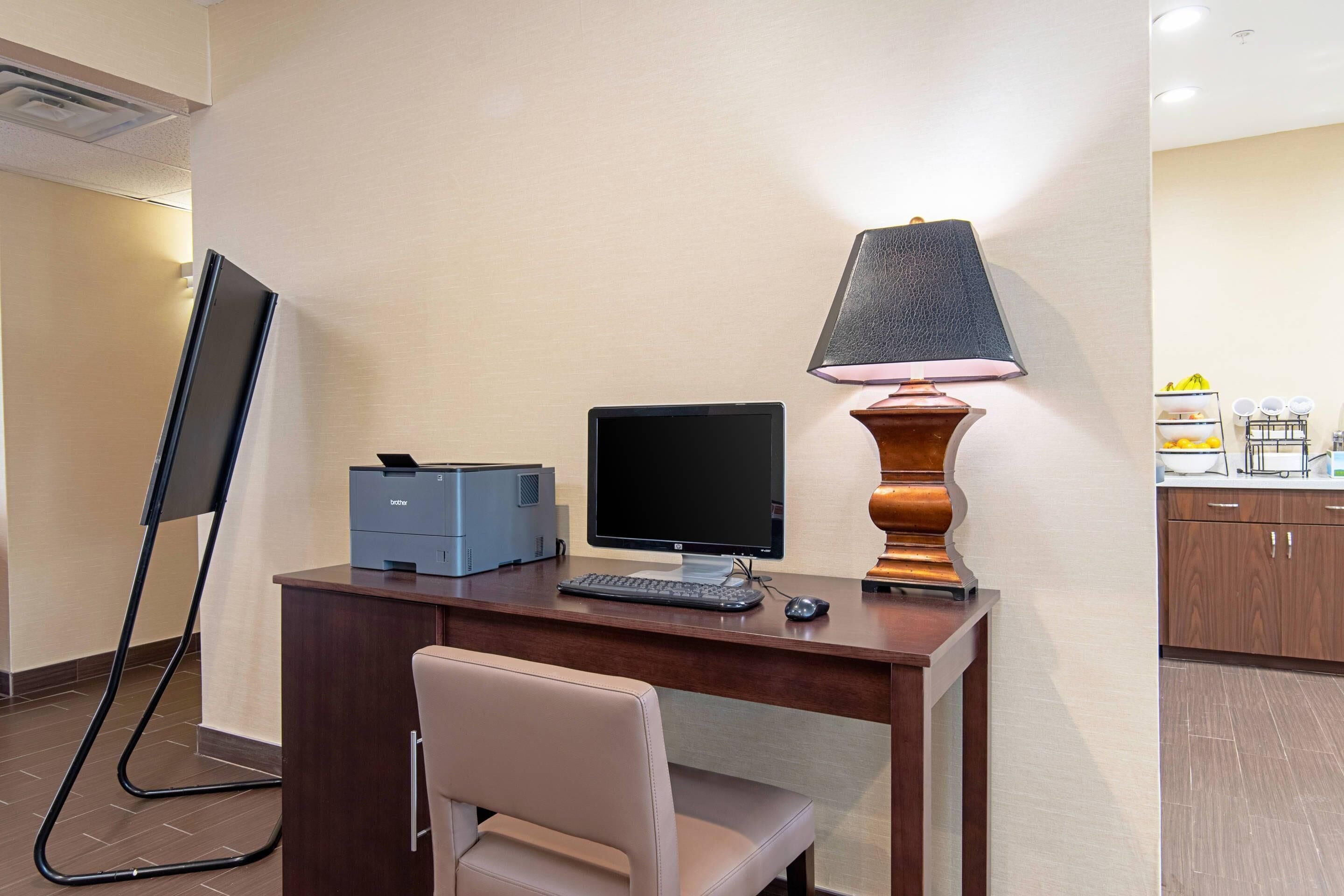 A hotel room with a desk, a computer, a printer, a lamp, a chair, and a TV.