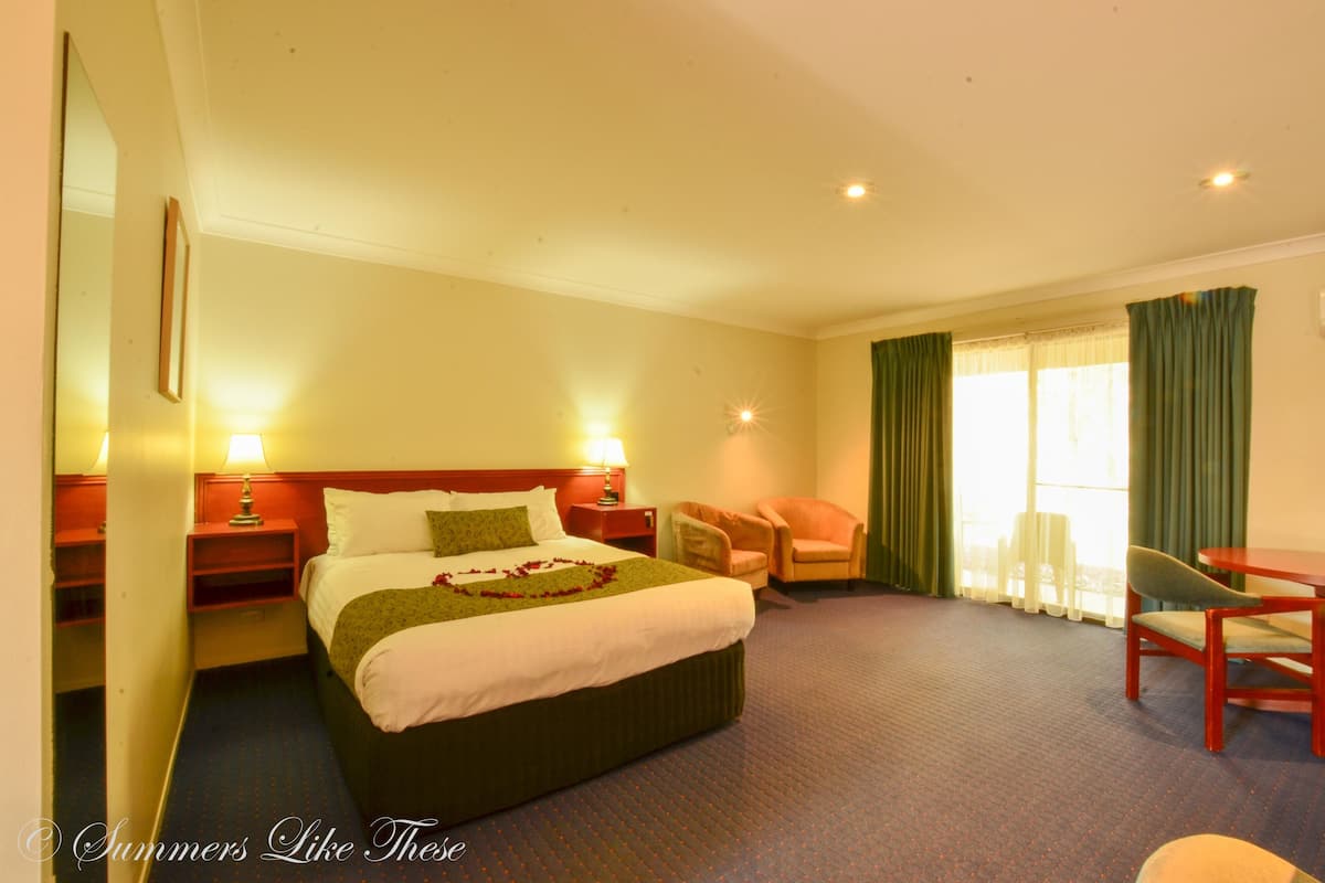 Habitación de hotel con una cama grande, dos mesitas de noche, una silla, una mesita pequeña y un ventanal con cortinas.