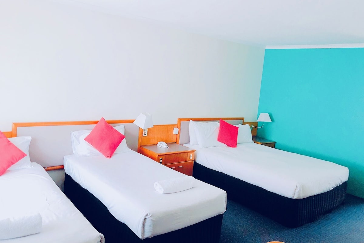 Habitación de hotel con tres camas individuales, cada una con ropa de cama blanca y almohadas rosadas. Hay mesitas de noche con lámparas y un teléfono. La habitación tiene una pared de acento color turquesa y suelo alfombrado.