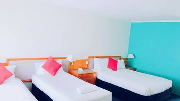 Habitación de hotel con tres camas individuales, cada una con ropa de cama blanca y almohadas rosadas. Hay mesitas de noche con lámparas y un teléfono. La habitación tiene una pared de acento color turquesa y suelo alfombrado.
