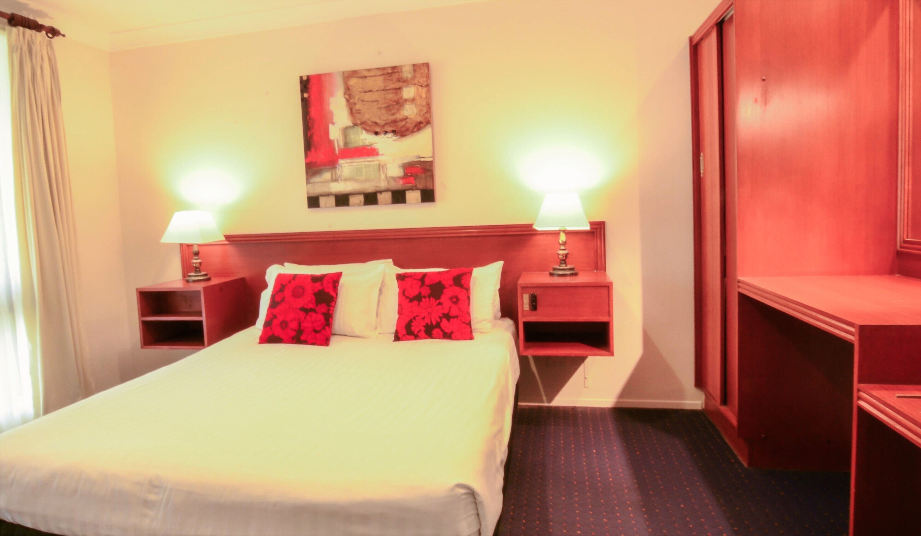 A hotel room with a large bed, two bedside tables with lamps, a wooden headboard, a painting on the wall, and a red wardrobe.