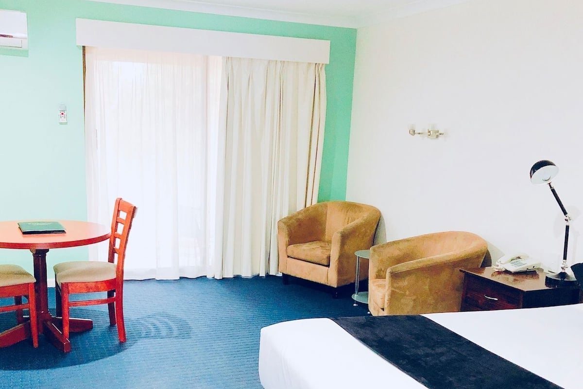 Una habitación de hotel con una cama, un escritorio con una silla, dos sillones, una ventana con cortinas y una mesita de noche con una lámpara.