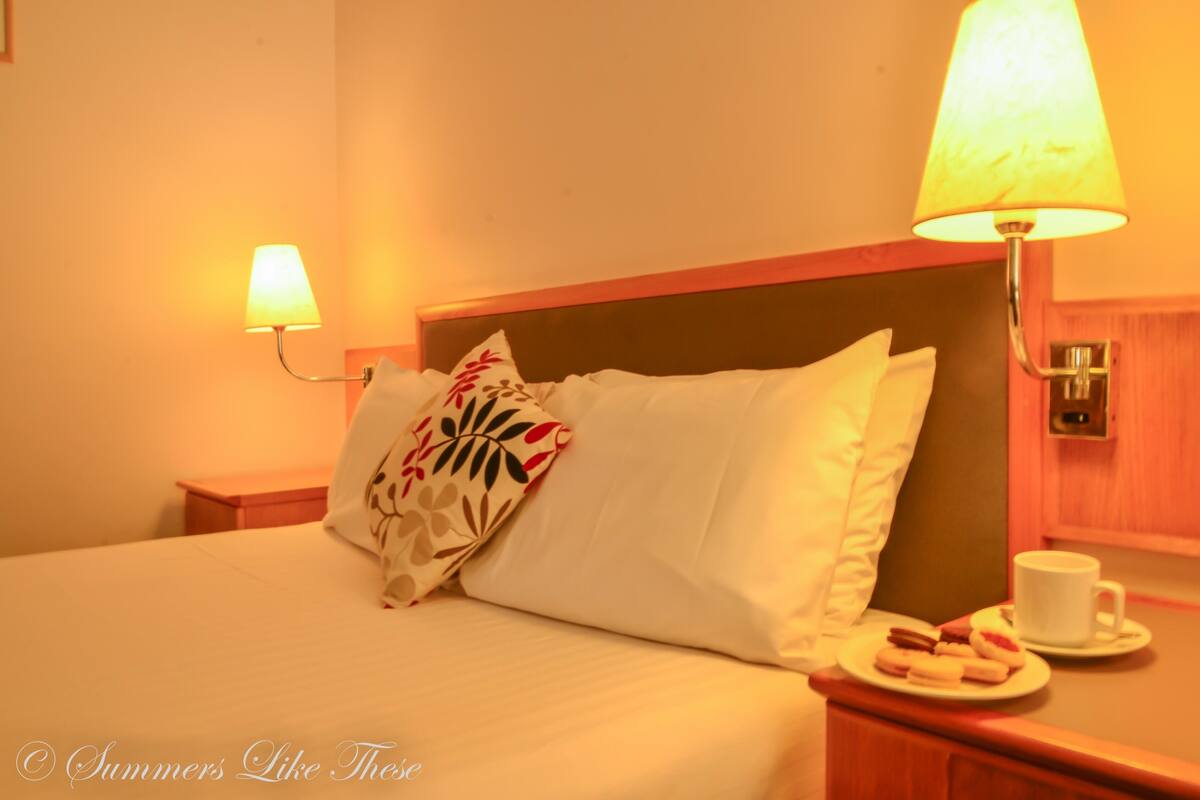 Habitación de hotel con cama, dos lámparas de noche, mesita de noche con una taza y un plato de comida, y cabecero de madera.