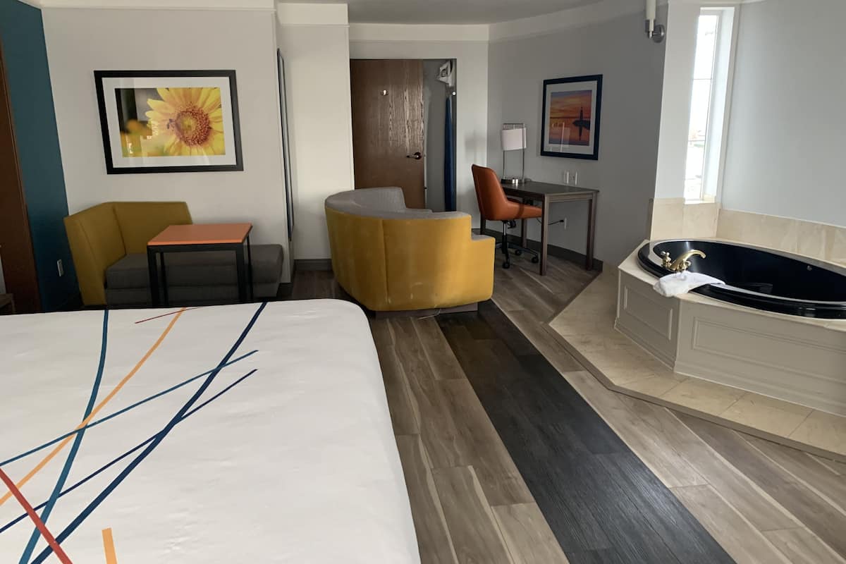 A hotel room with a bed, a desk, a chair, a sofa, a bathroom with a bathtub, and artwork on the walls.