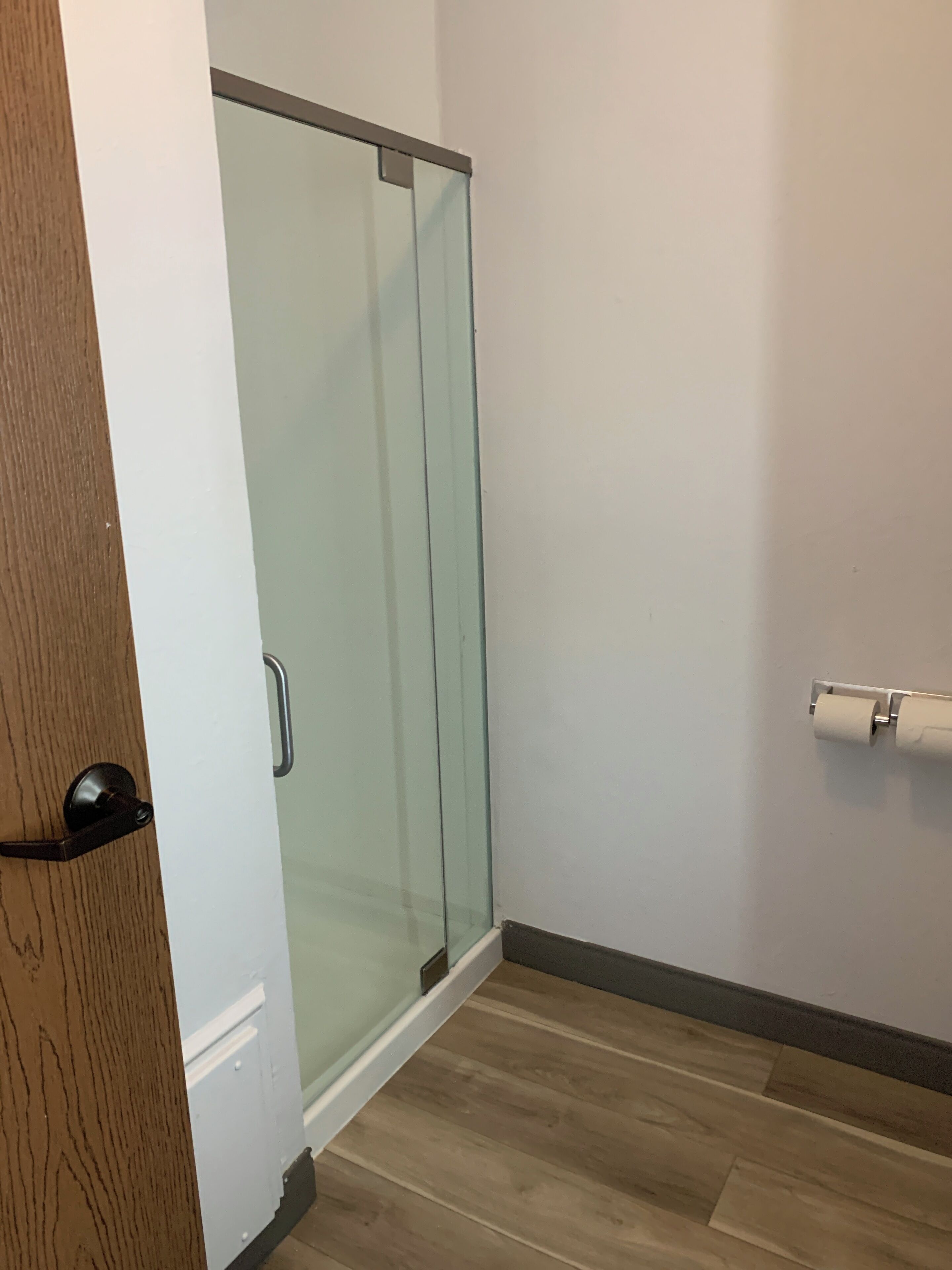 Un baño con mampara de vidrio, dispensador de papel higiénico y piso de madera.