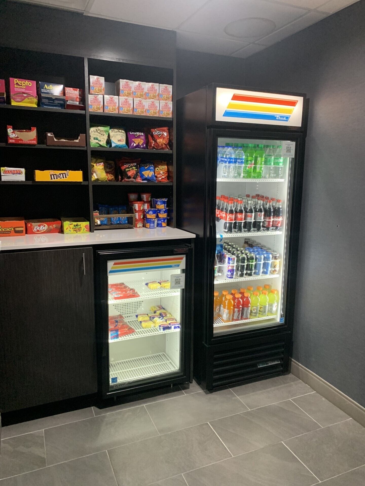 Un petit présentoir réfrigéré contenant diverses boissons et collations, notamment des sodas et des chips, dans un point de vente.