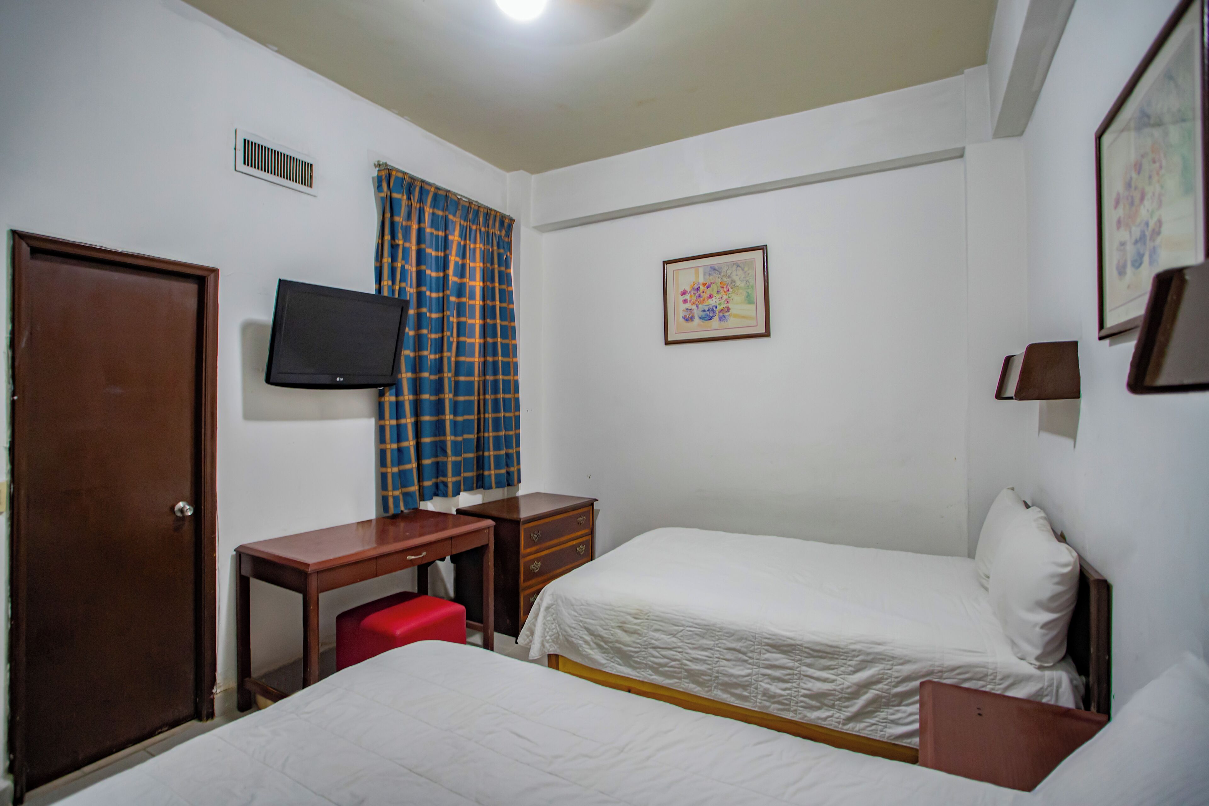 A hotel room with a bed, a desk with a chair, a television, a door, and a window with curtains.