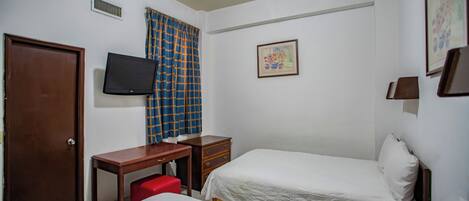 A hotel room with a bed, a desk with a chair, a television, a door, and a window with curtains.