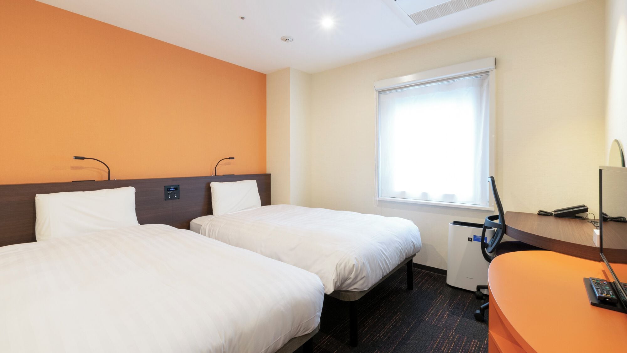 A hotel room with two beds, a desk, a chair, a window with blinds, and a control panel on the wall.