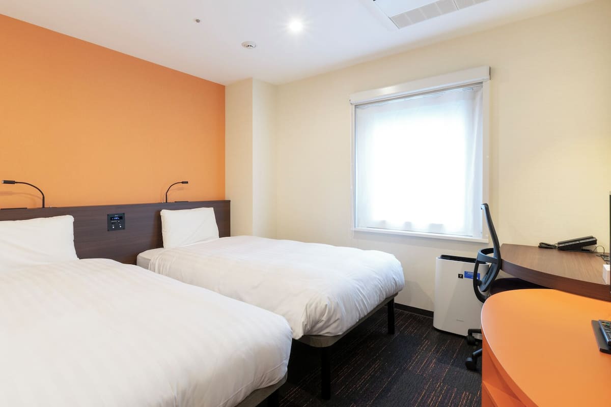 A hotel room with two beds, a desk, a chair, a window with blinds, and a control panel on the wall.