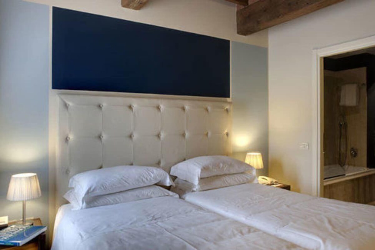 Una cama doble con cabecera capitonada, dos lámparas de noche y una mesita de noche con un libro.