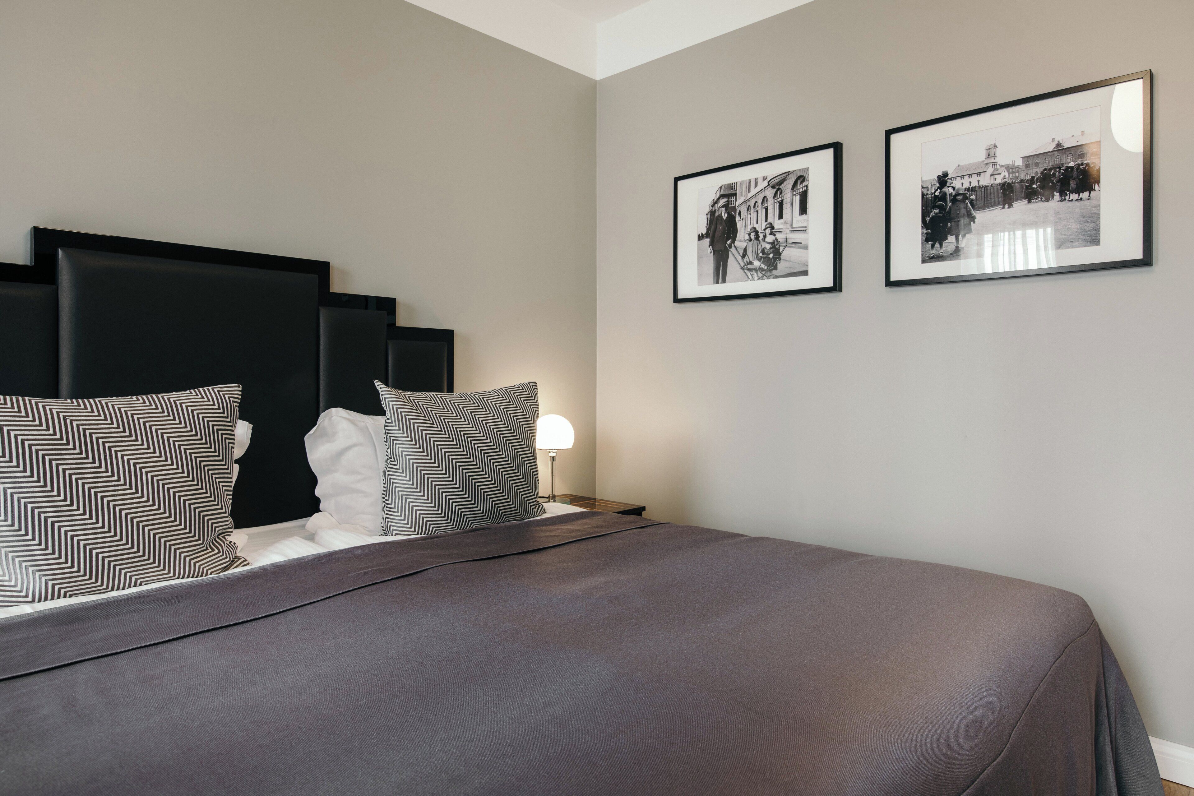 A neatly made bed with a grey bedspread and patterned pillows, a bedside lamp, and two framed black and white photographs on the wall.