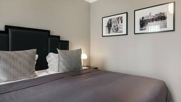 A neatly made bed with a grey bedspread and patterned pillows, a bedside lamp, and two framed black and white photographs on the wall.