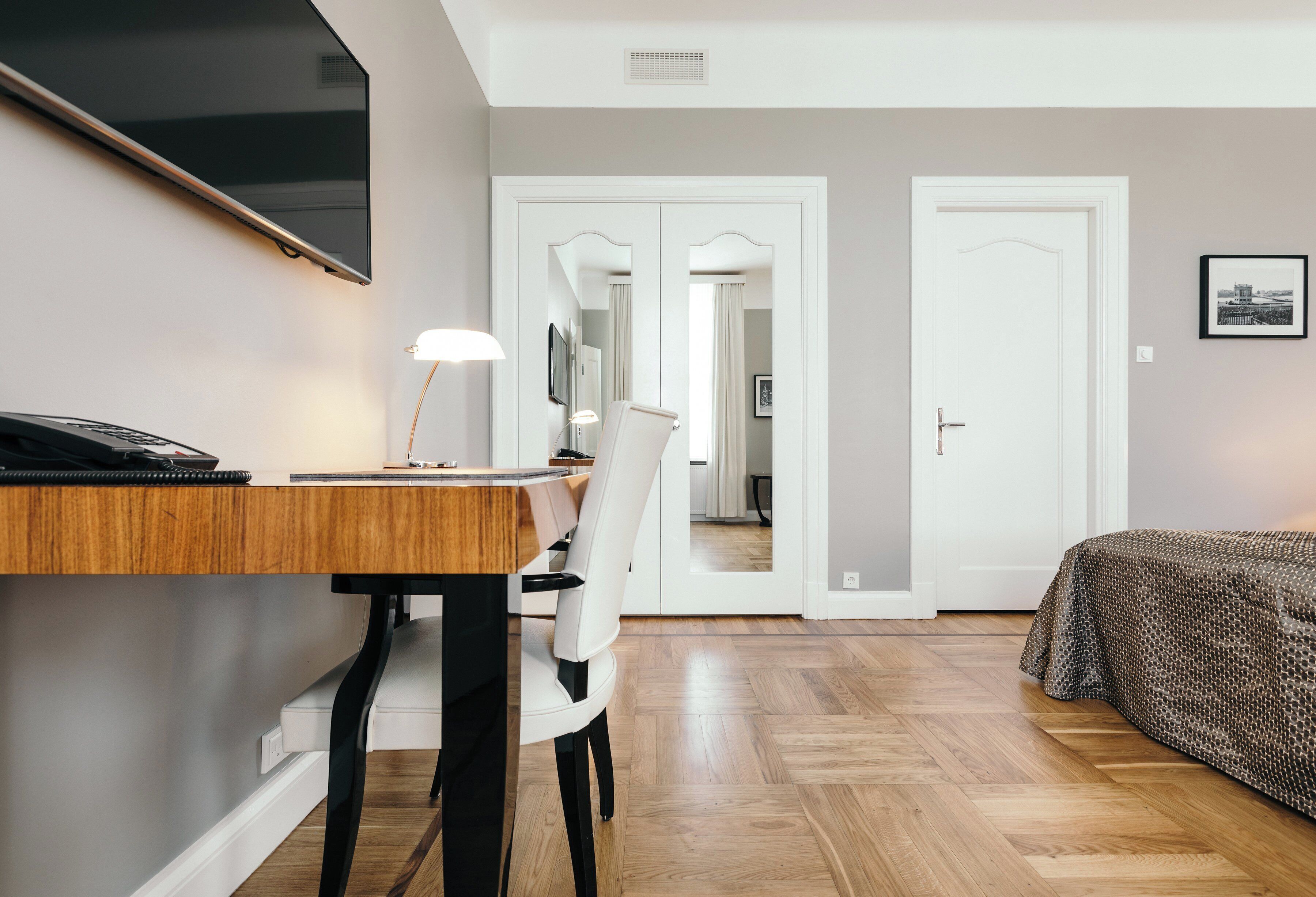 Une chambre d’hôtel avec un bureau en bois, une chaise, un lit avec une literie à motifs, un téléviseur à écran plat fixé au mur et une porte donnant sur une autre pièce.