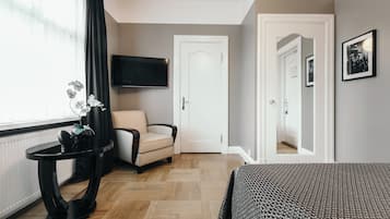 Una habitación de hotel con una cama, una silla, una mesita con un florero, un televisor montado en la pared y una puerta que da a otra habitación.