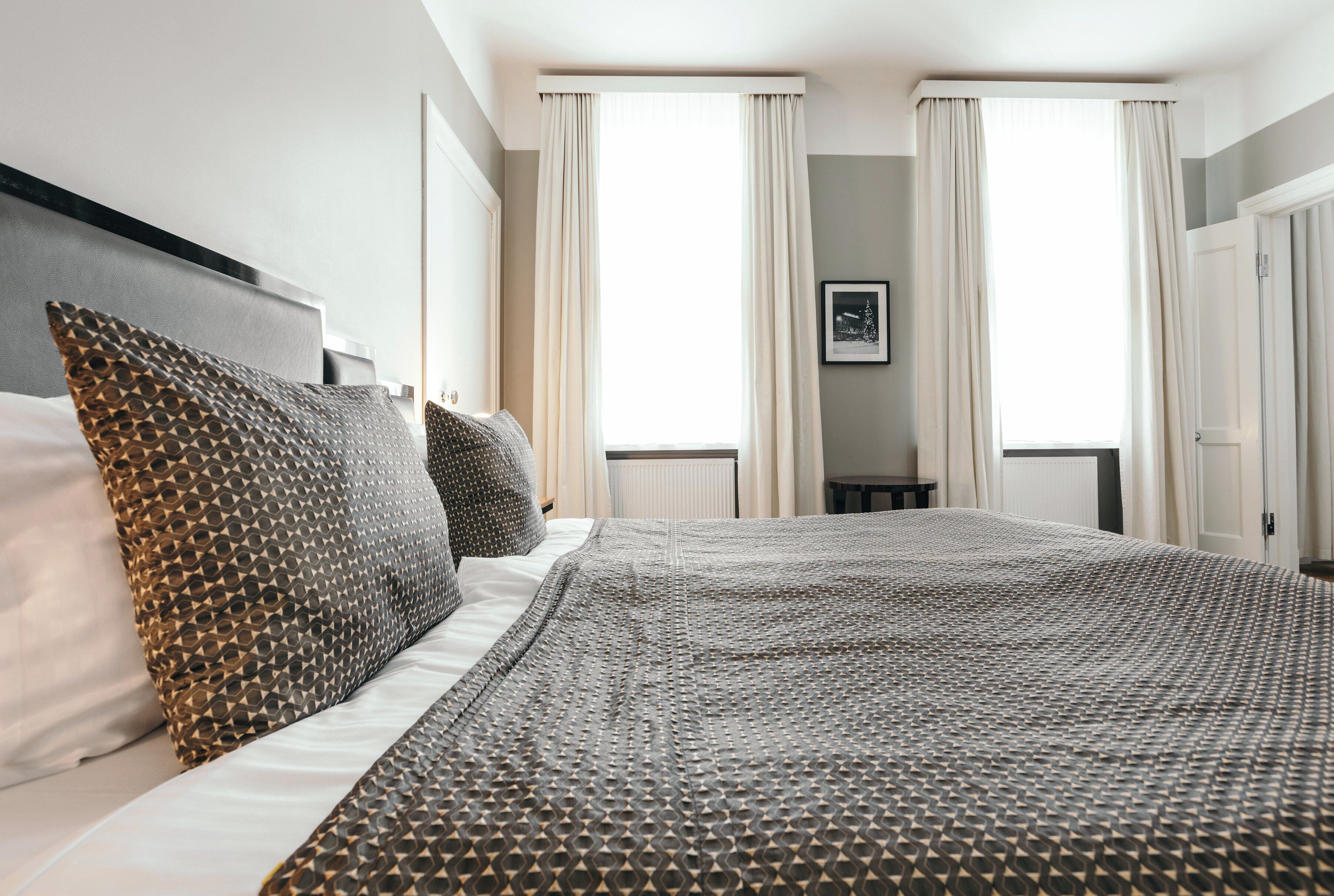 A neatly made bed with patterned bedding, a headboard, and two windows with curtains.