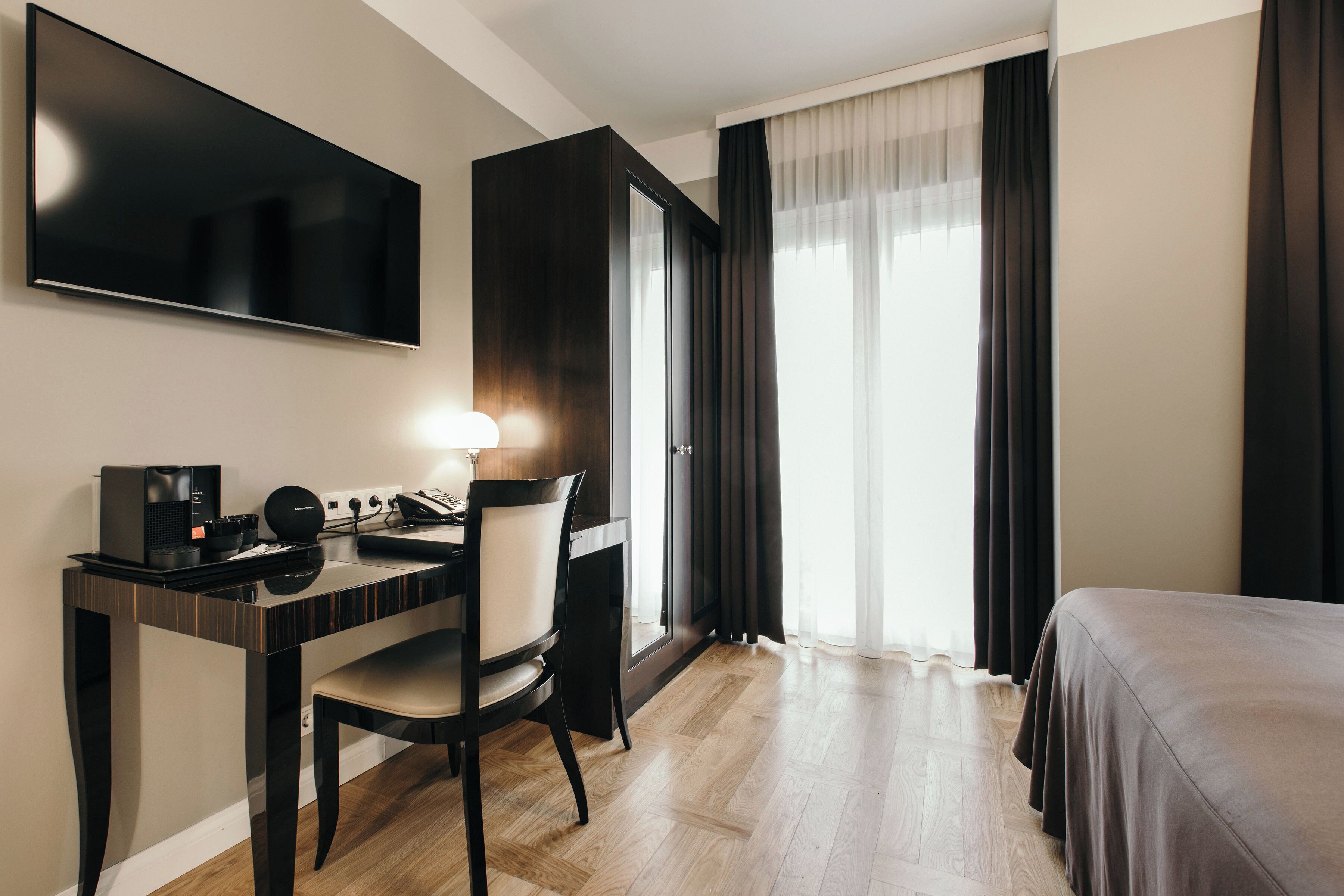 A hotel room with a bed, a desk with a computer, a chair, and a TV mounted on the wall.