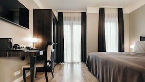 Habitació d'hotel amb llit, escriptori amb cadira, televisió i finestra gran amb cortines.